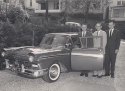 Three men stand beside a vintage car with the driver’s door open in a driveway.