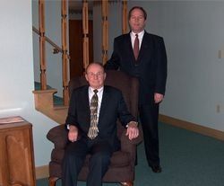 Two men in dark suits, one seated and one standing, in a carpeted office hallway.