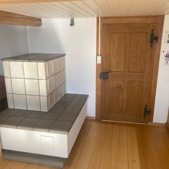 Ein weiß gekachelter Holzofen steht neben einer Holztür in einem Zimmer mit hellem Holzboden.