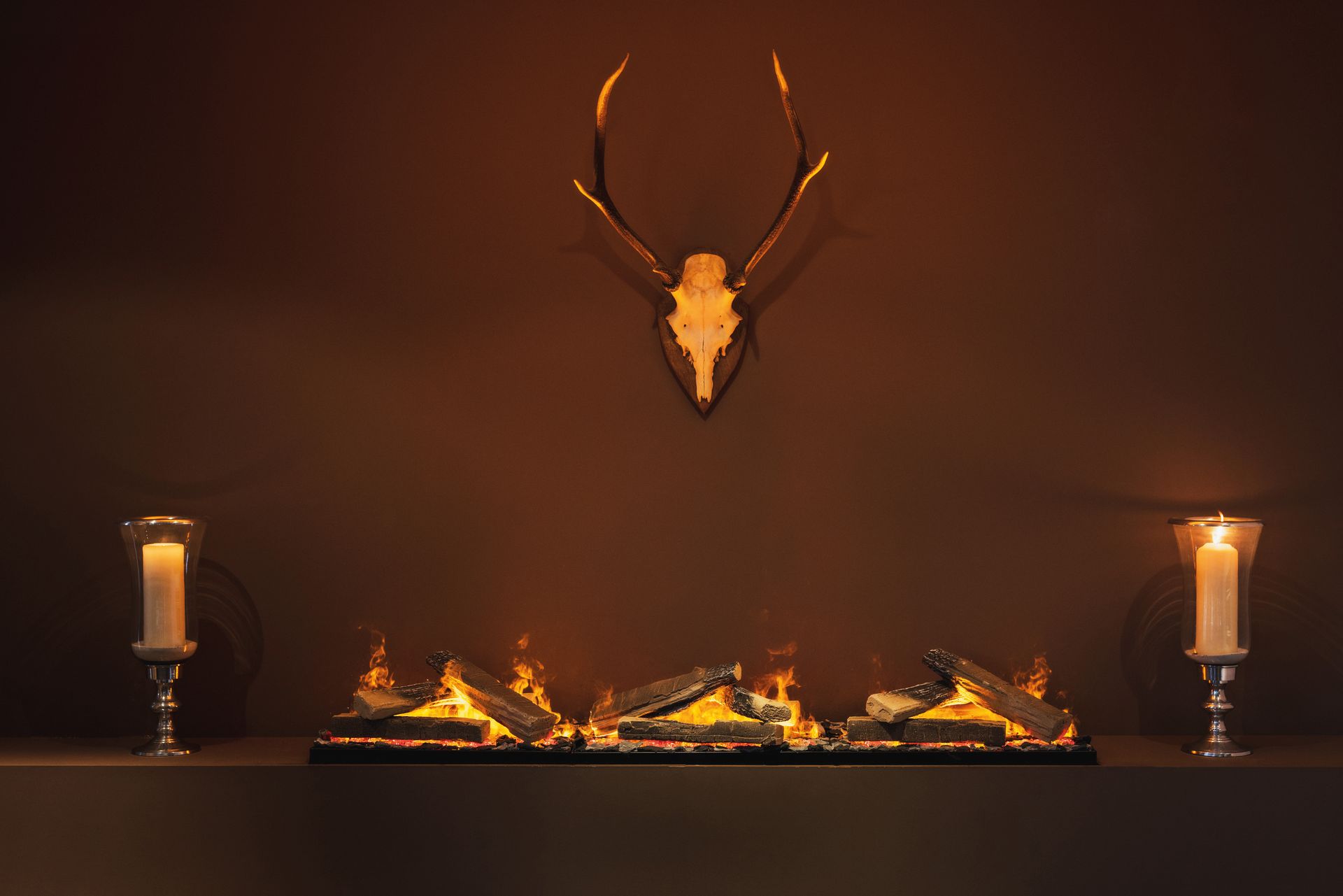 Kamin mit lodernden Flammen, flankiert von Kerzen, darüber ein Hirschschädel.
