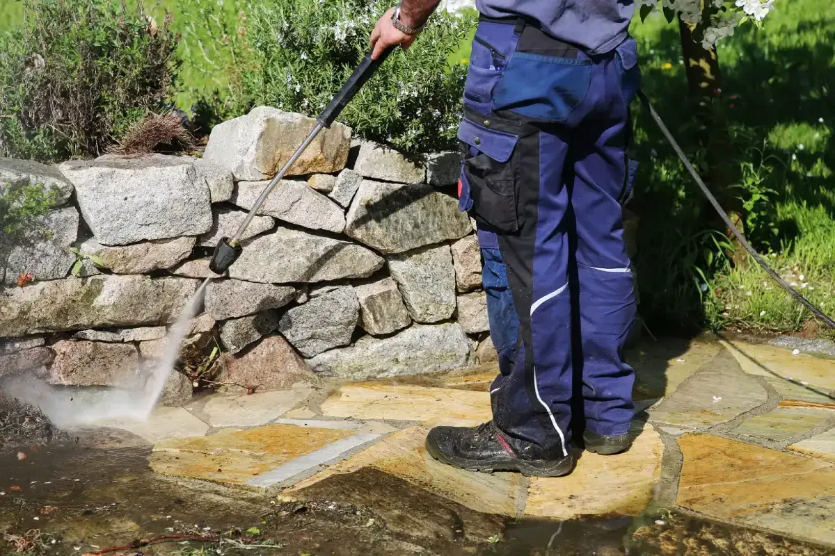Person pressure-washing a stone patio beside a rock wall with a black hose.