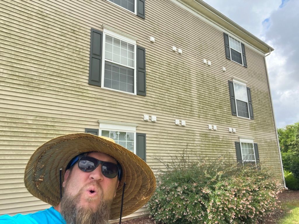 Man in straw hat and sunglasses taking a selfie in front of a tan apartment building with black shutters