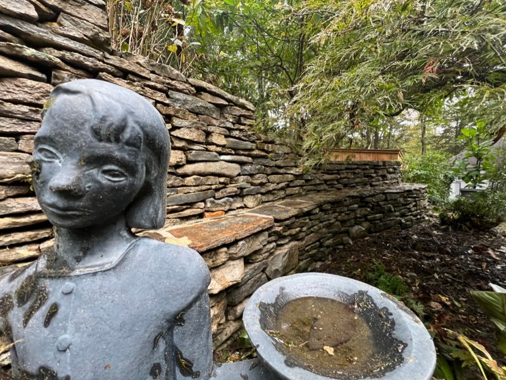 Bronze child statue beside a stone wall and shallow basin in a leafy garden