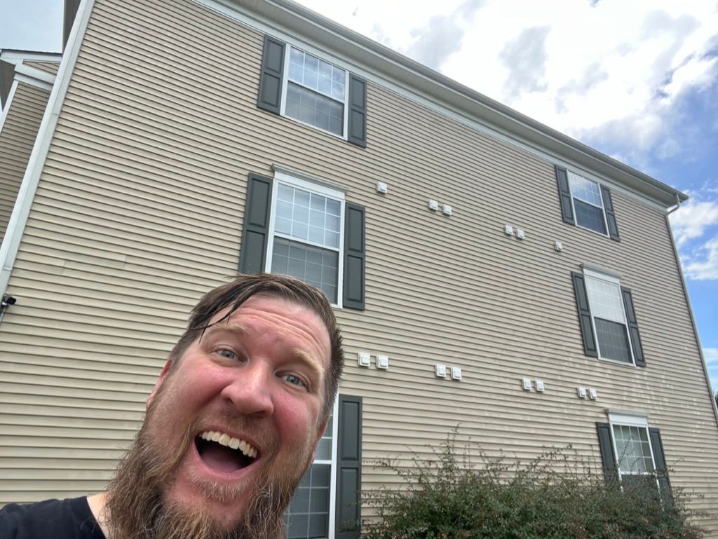 Smiling bearded man selfie in front of a tan apartment building under a cloudy sky
