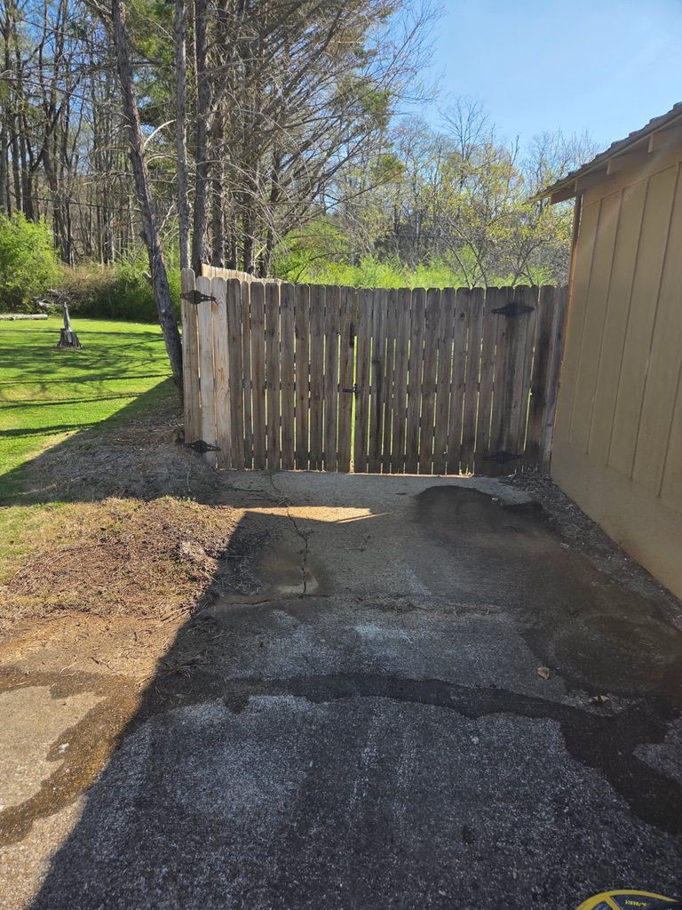 Backyard view with cracked pavement, weathered wooden gate, and beige shed beside a grassy yard.