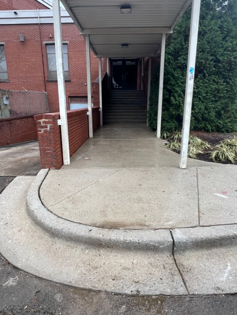 Covered sidewalk ramp leading to stairs at a brick building entrance, with concrete curb and hedges nearby