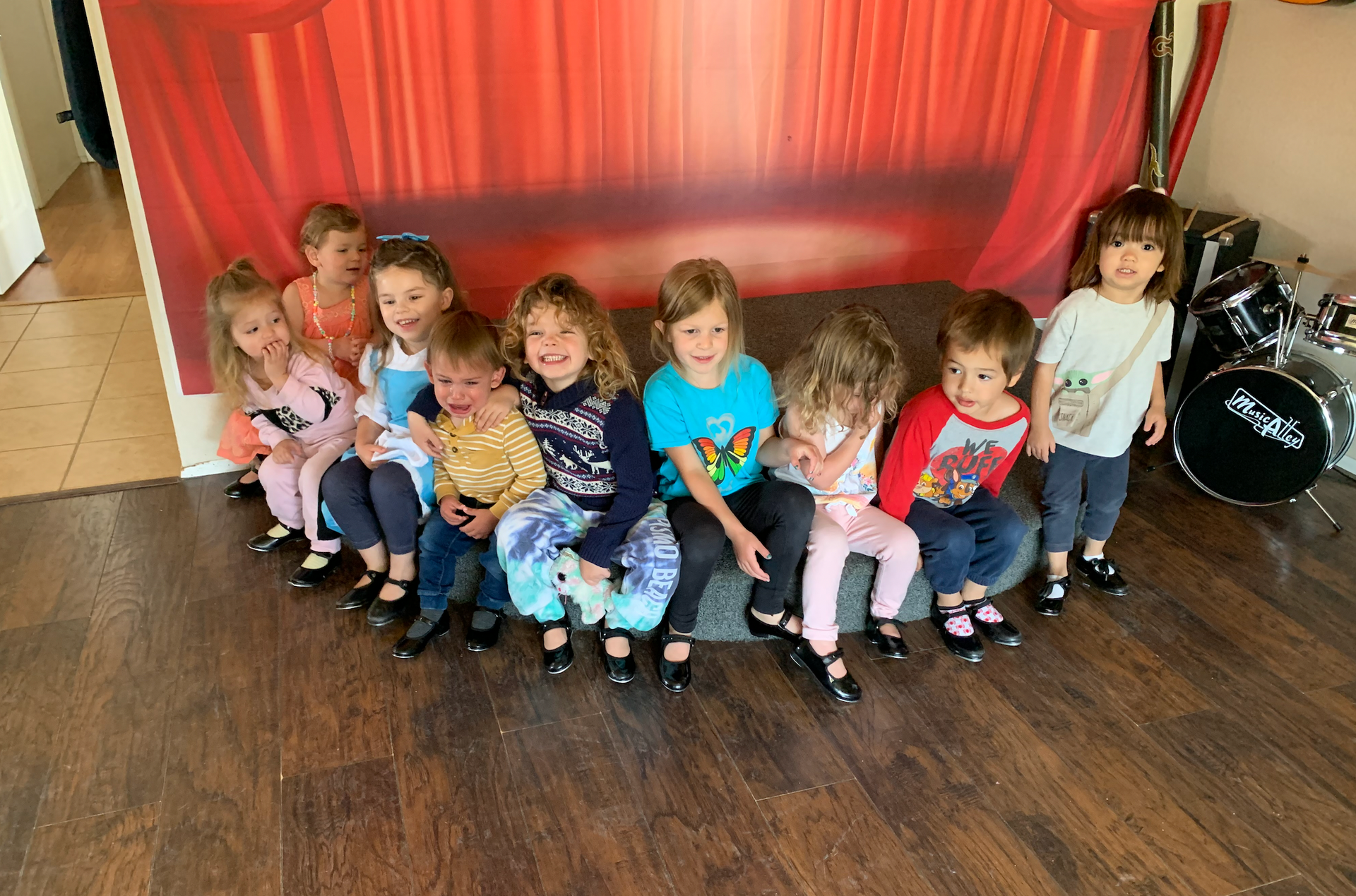A group of children are sitting on the floor in front of a red curtain.