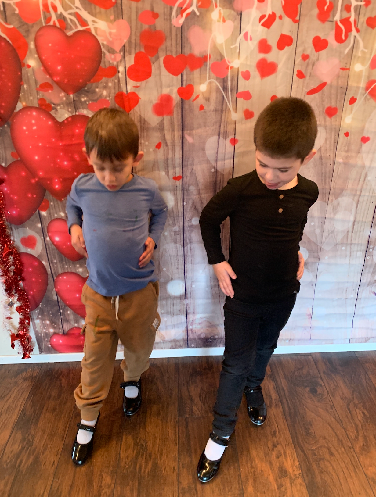 Two young boys are standing in front of a wooden wall with hearts on it.