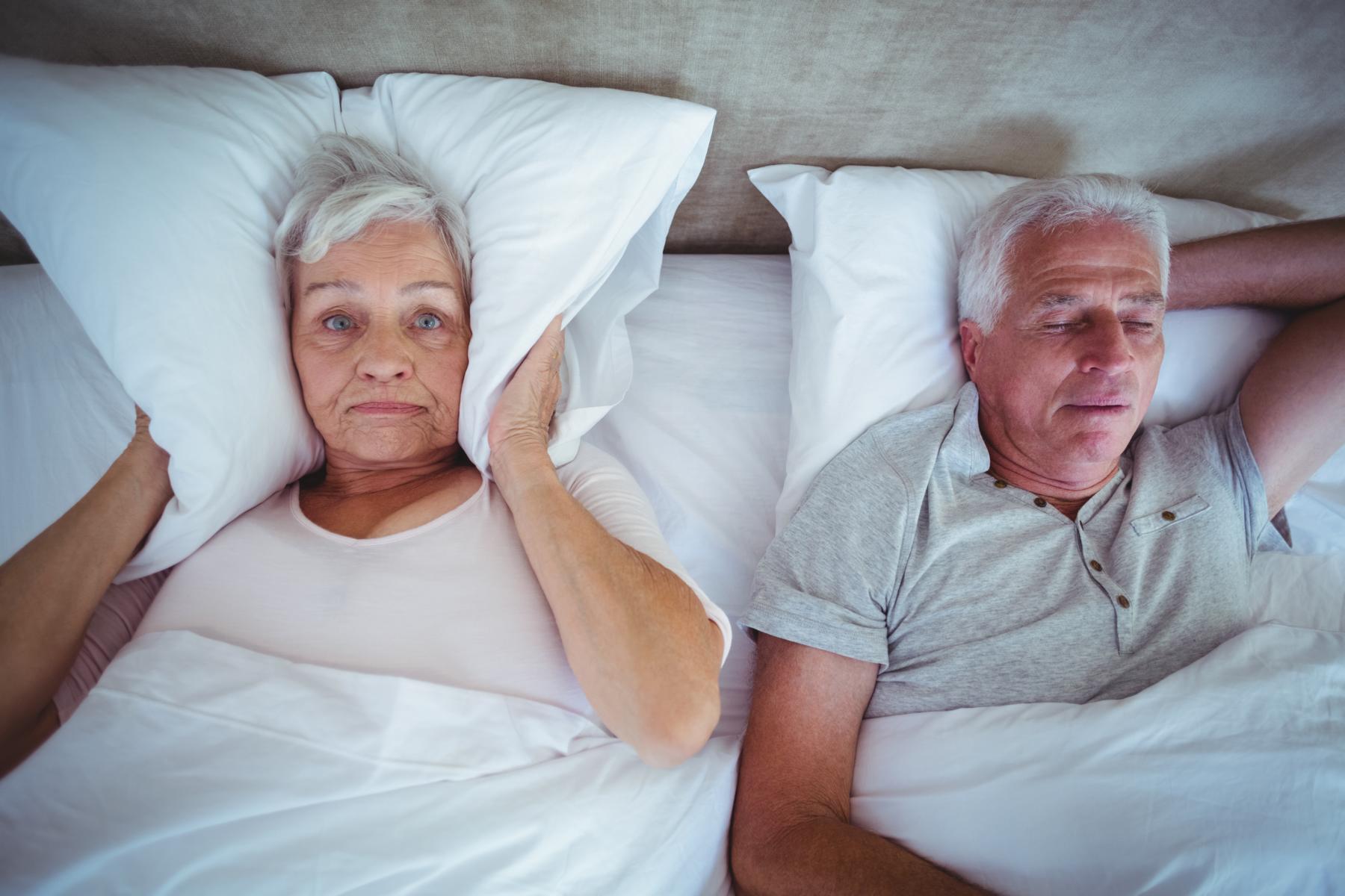 Ein Mann und eine Frau liegen im Bett und halten sich Kissen die Ohren zu.