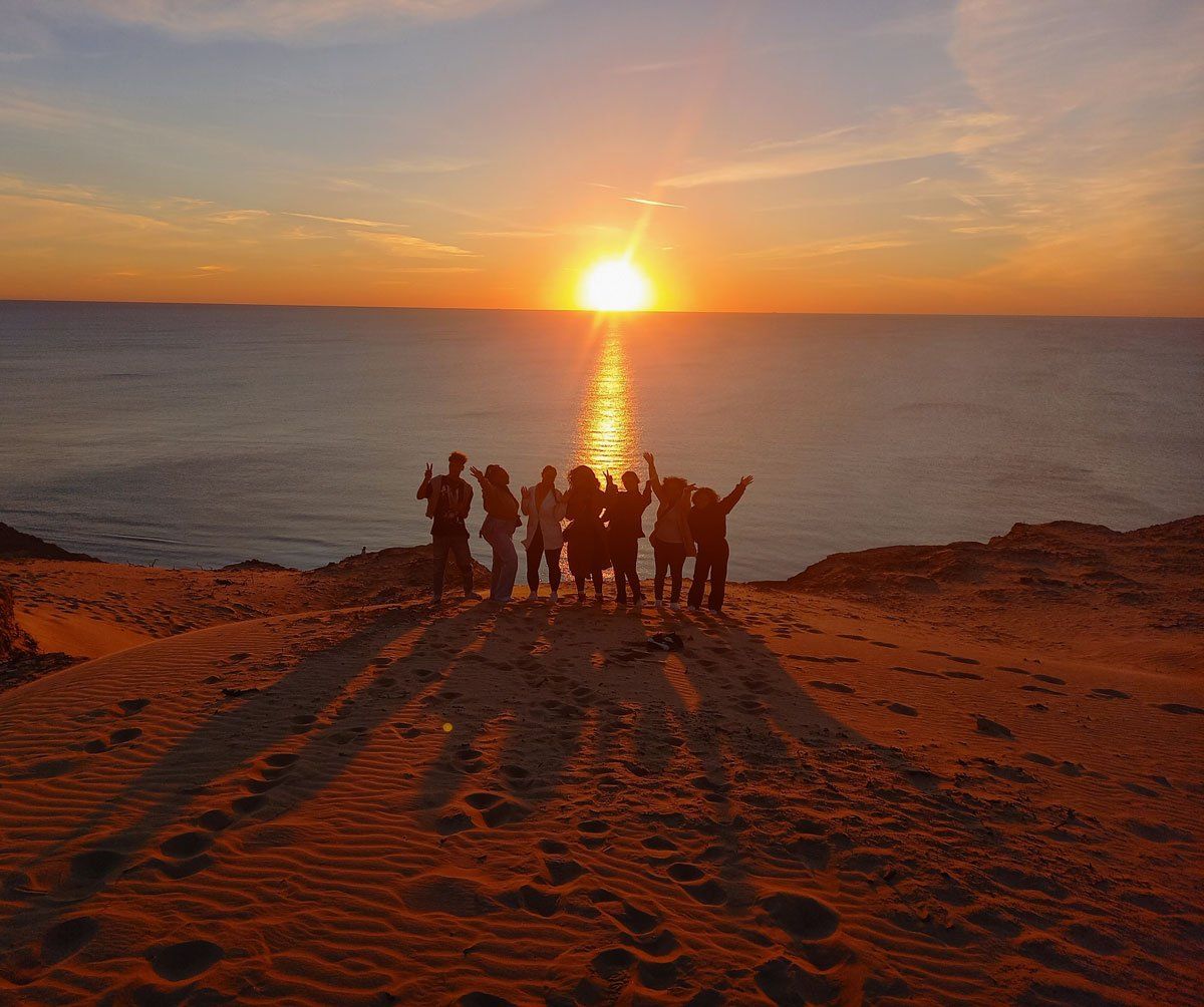 Eine Gruppe von Menschen steht auf einer Sanddüne und beobachtet den Sonnenuntergang über dem Meer