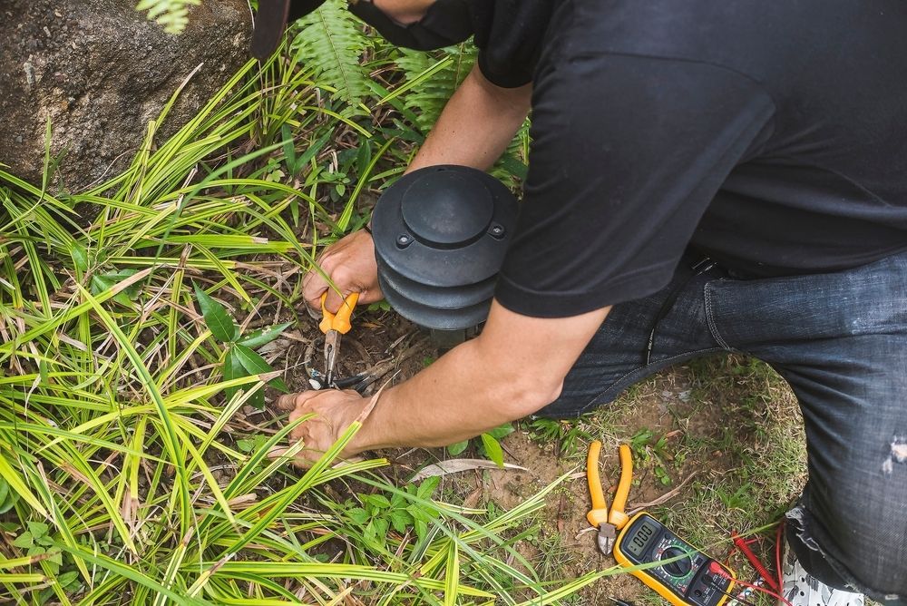 Person in Black Shirt and Jeans Using Tools on Wires Next to A Light Fixture — Lads Lawns & Gardens in Bentley Park, QLD