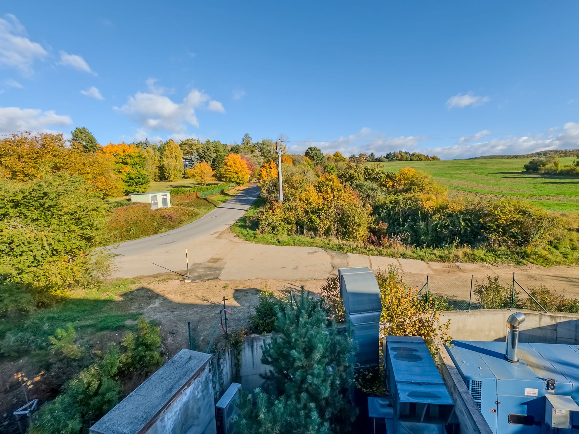 A sunny, autumnal landscape featuring a winding road bordered by colorful trees, greenery, and blue industrial equipment.