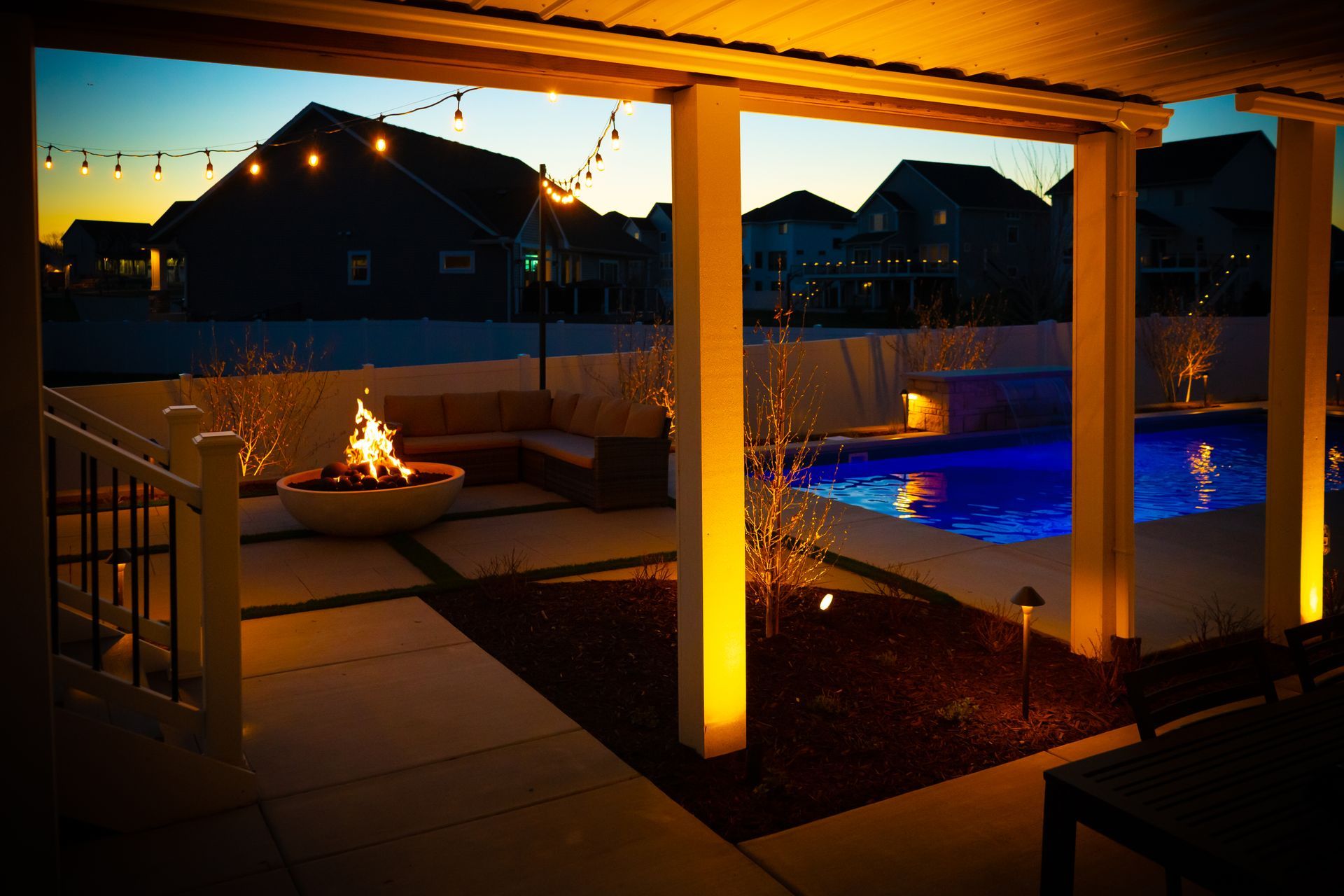 Backyard patio at dusk with a fire pit, lit pergola posts, and a glowing blue pool