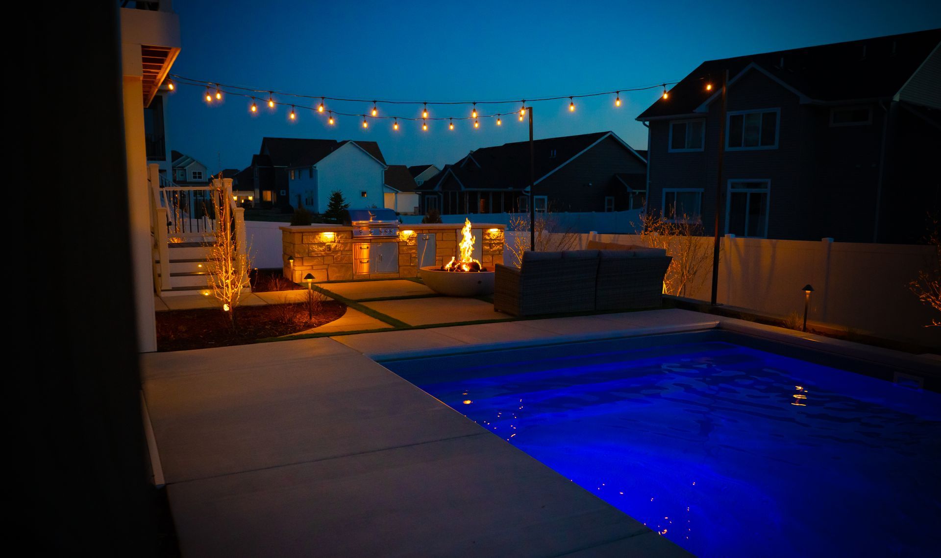 Backyard at dusk with lit pool, patio lights, and neighboring houses in the background