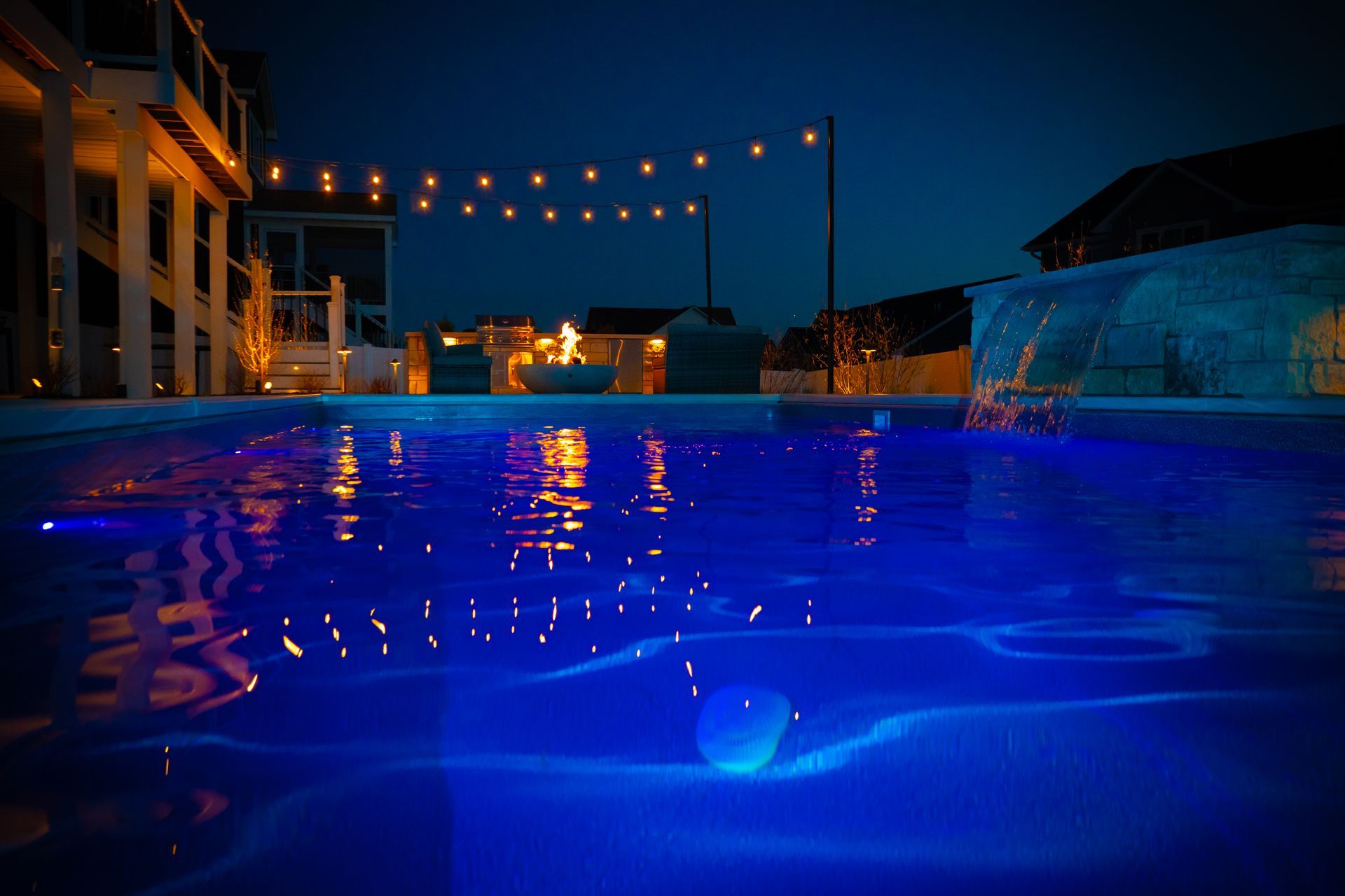 Blue-lit swimming pool at night with string lights overhead and building reflections on the water