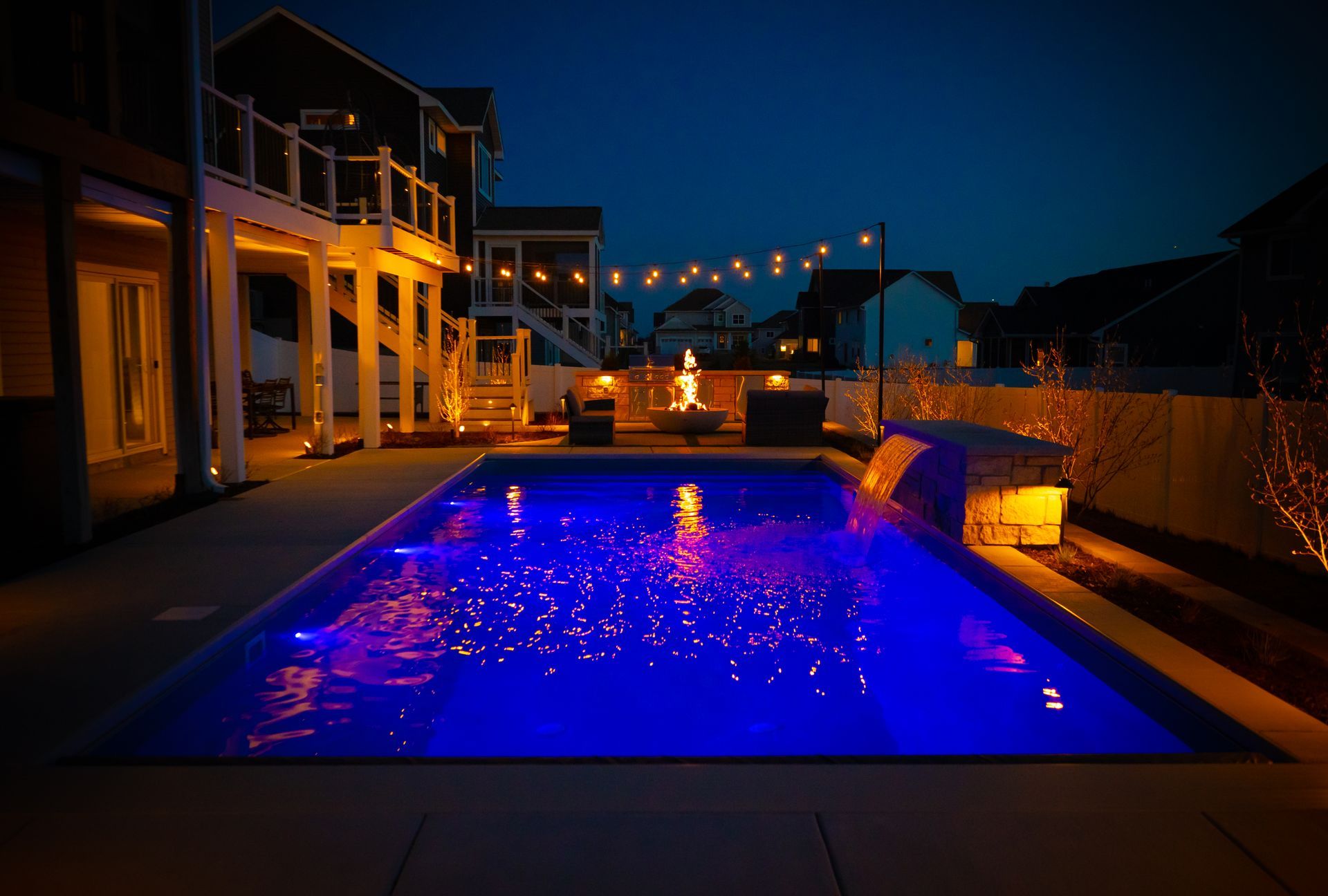 Illuminated backyard pool at night beside a house, glowing blue with string lights in the background