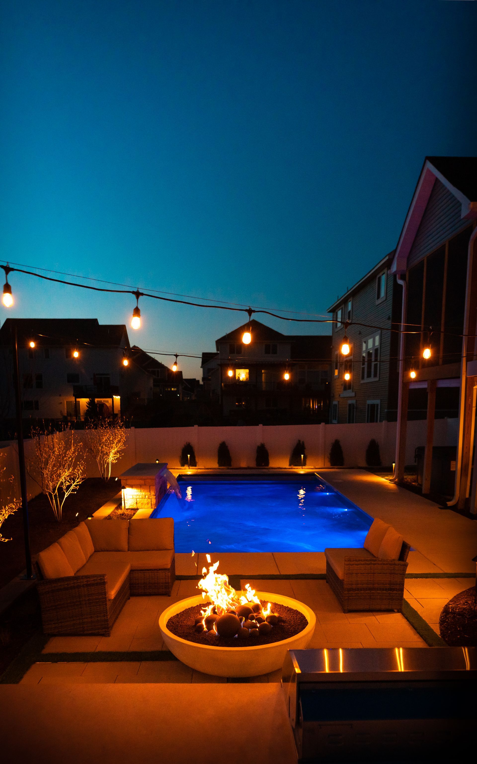 Rooftop pool deck at dusk with string lights, lounge chairs, and a glowing fire pit near the water