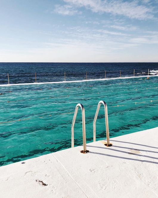 Piscina all'aperto con scaletta, acqua turchese, vista sull'oceano, cielo azzurro e luce solare intensa.