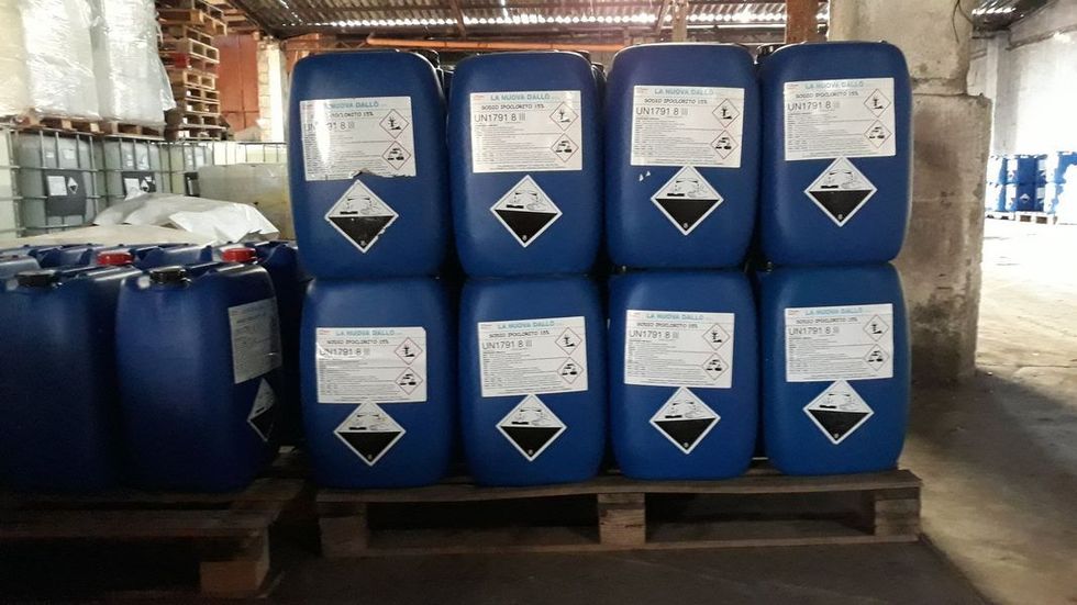Barili blu impilati su pallet, ognuno etichettato con simboli di pericolo, in un ambiente di magazzino.