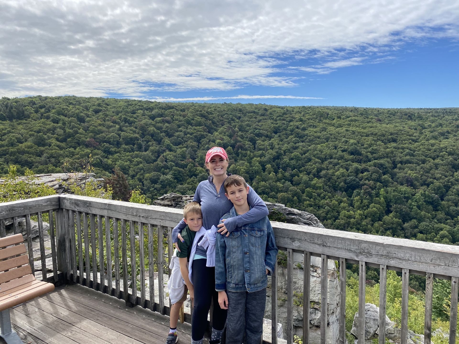 Woman and two boys on a wooden overlook, hugging, overlooking lush green forest under a blue sky with clouds. Breathe In, Hike Out: The Best Local Trails for a Summer Escape