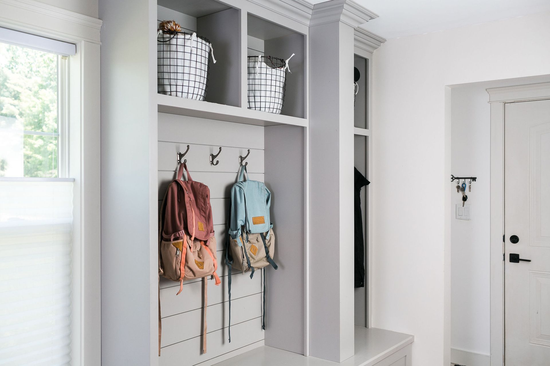 Mudroom with built-in storage, hooks holding backpacks, and baskets above. Bright and airy space, painted white and gray. Back-to-School, Back to Routine: Tips for Resetting After Summer