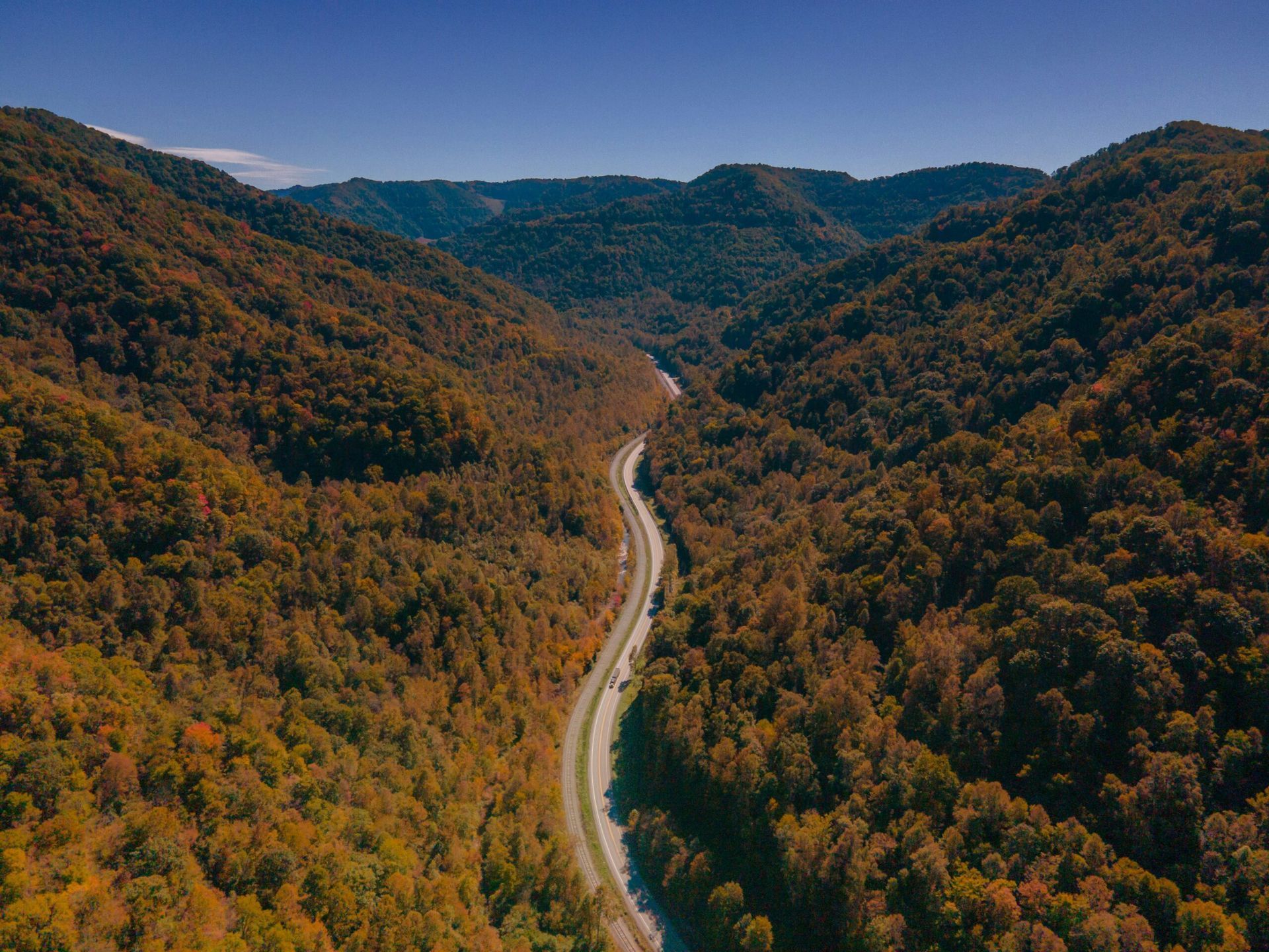 A woman is carrying a child on her back in a park. An aerial view of a road in the mountains surrounded by trees.