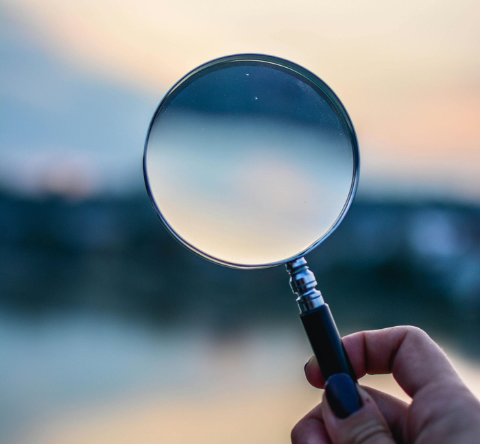 A hand holds a magnifying glass against a soft-focus, sunset-colored horizon.