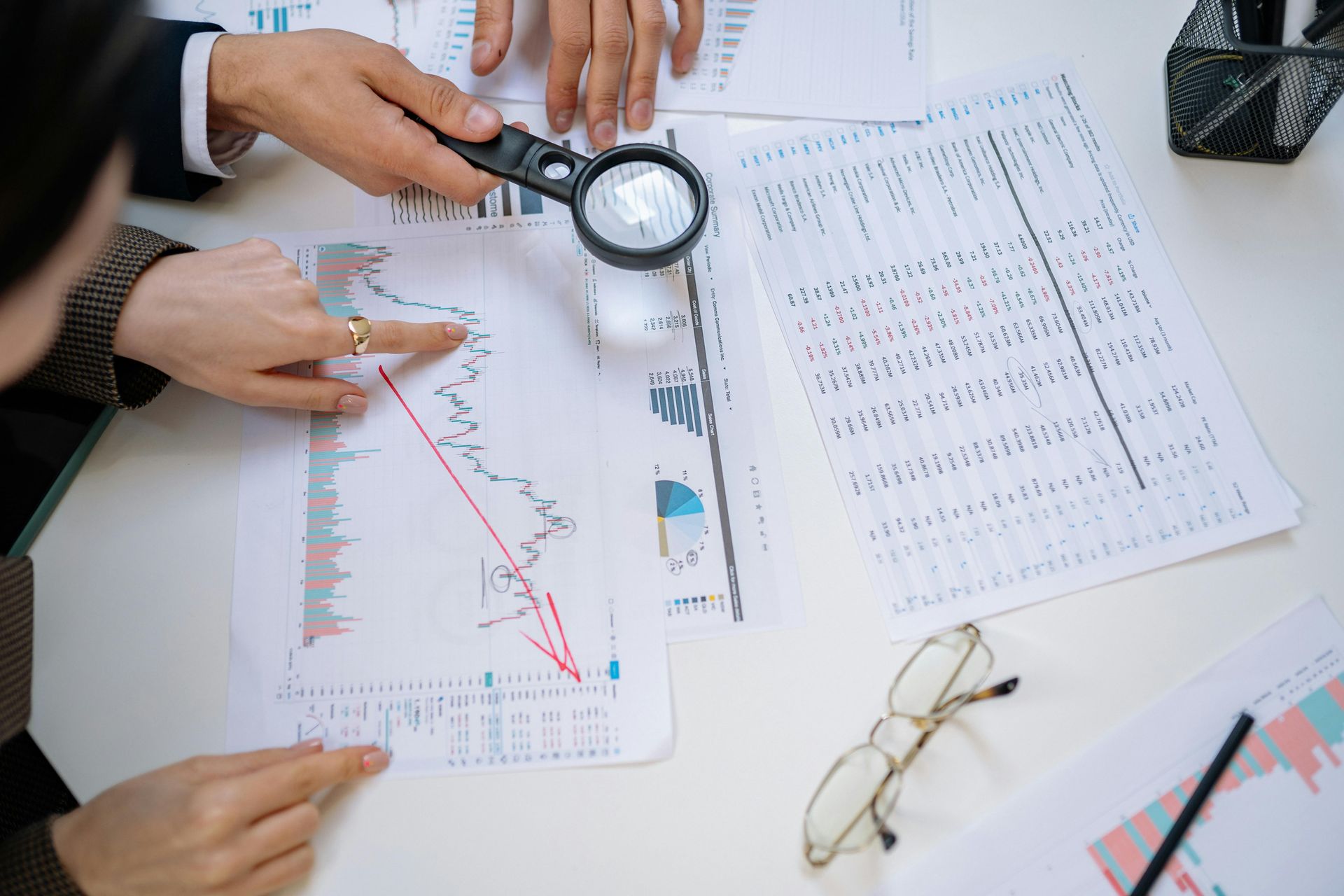 Two people analyze a printed line graph showing a downward trend, using a magnifying glass on a document-covered desk.