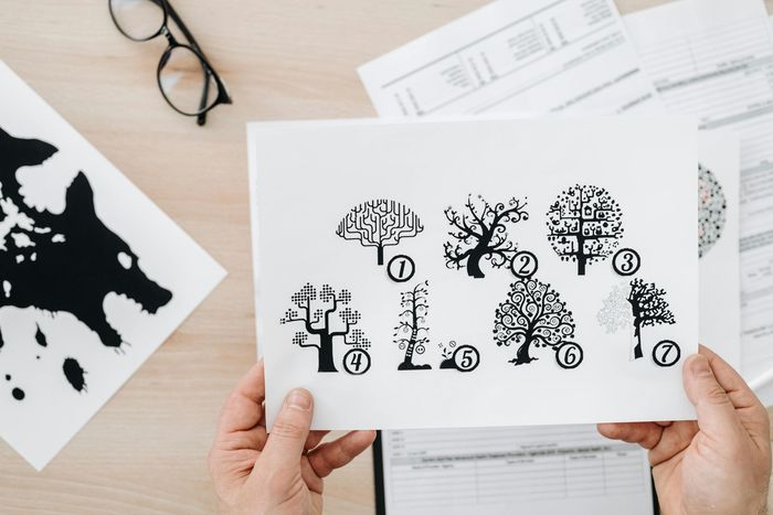 Hands hold a sheet displaying seven numbered tree illustrations above a desk with documents and an inkblot drawing.