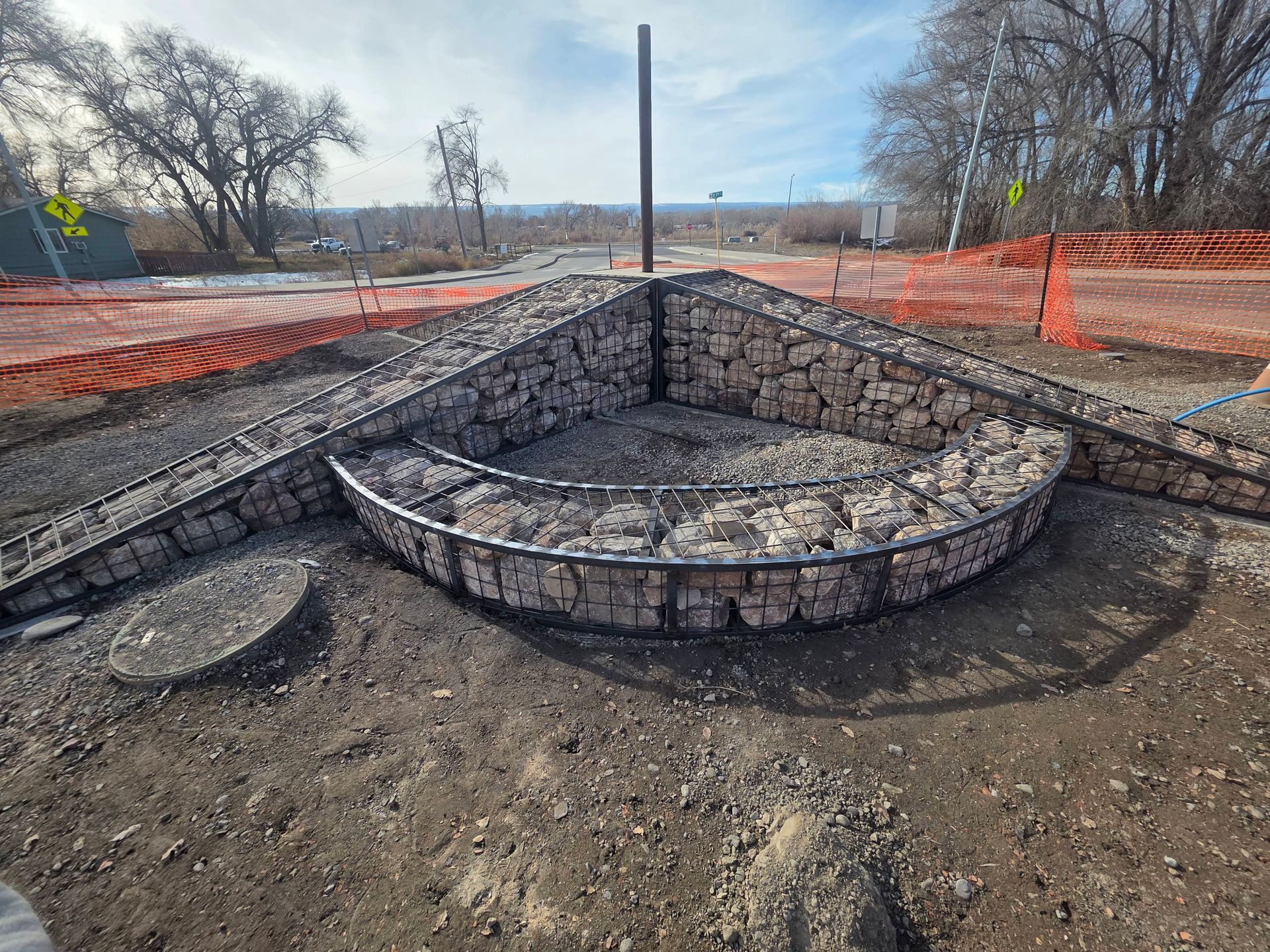 Stone fire pit under construction in a dirt lot, with orange safety fencing and a metal post.