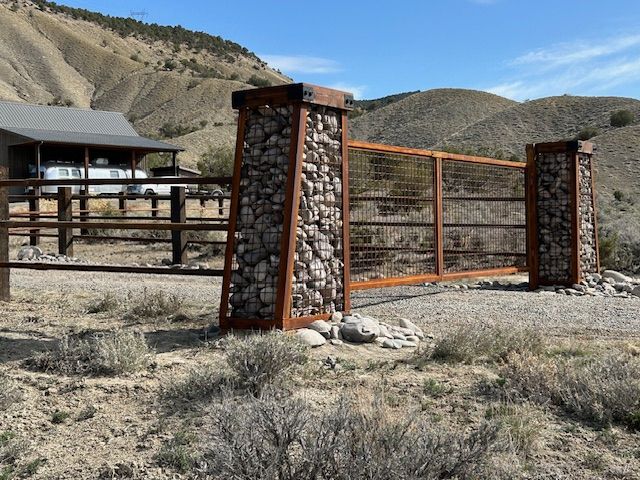 Rural ranch gate with stone pillars and rusty metal fencing in a dry, hilly landscape.