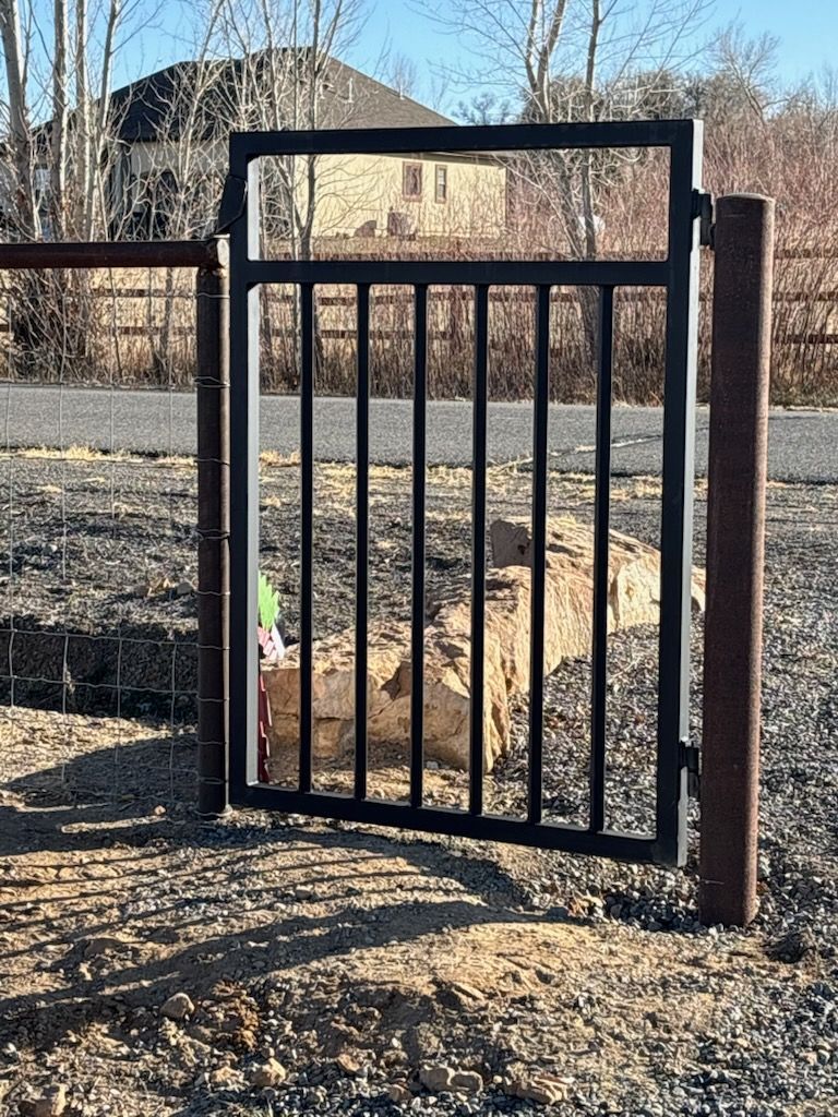 Black metal pedestrian gate beside a dirt road, with a house and leafless trees in the background