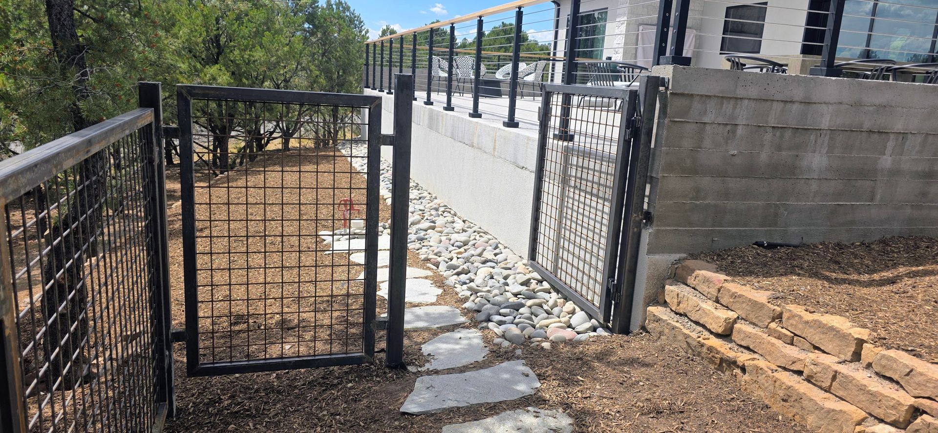 Black metal gate in a gravel-and-concrete yard beside a stone wall and fenced walkway under a blue sky