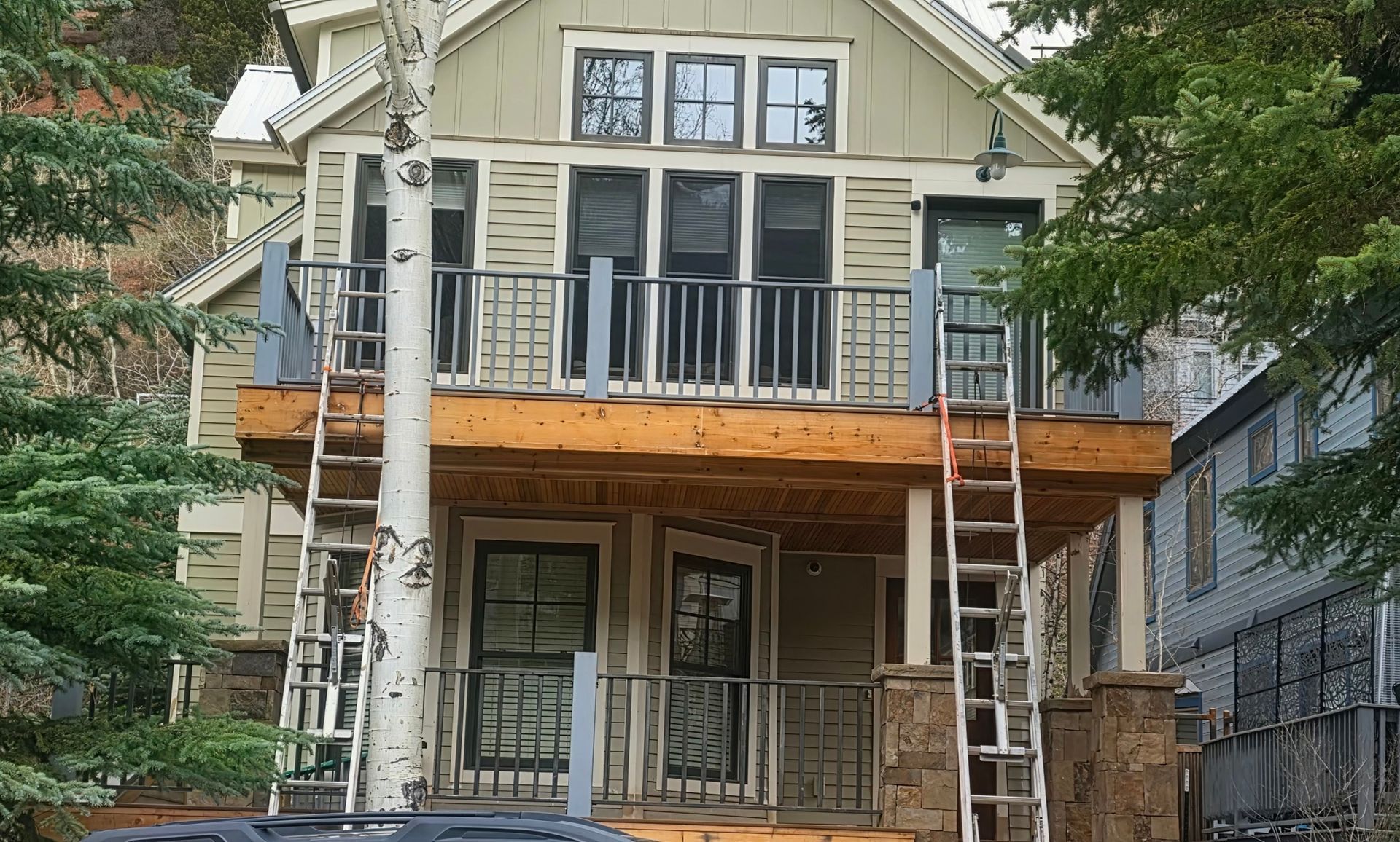 Two-story beige house with a front porch, balcony, and wooden siding, surrounded by trees