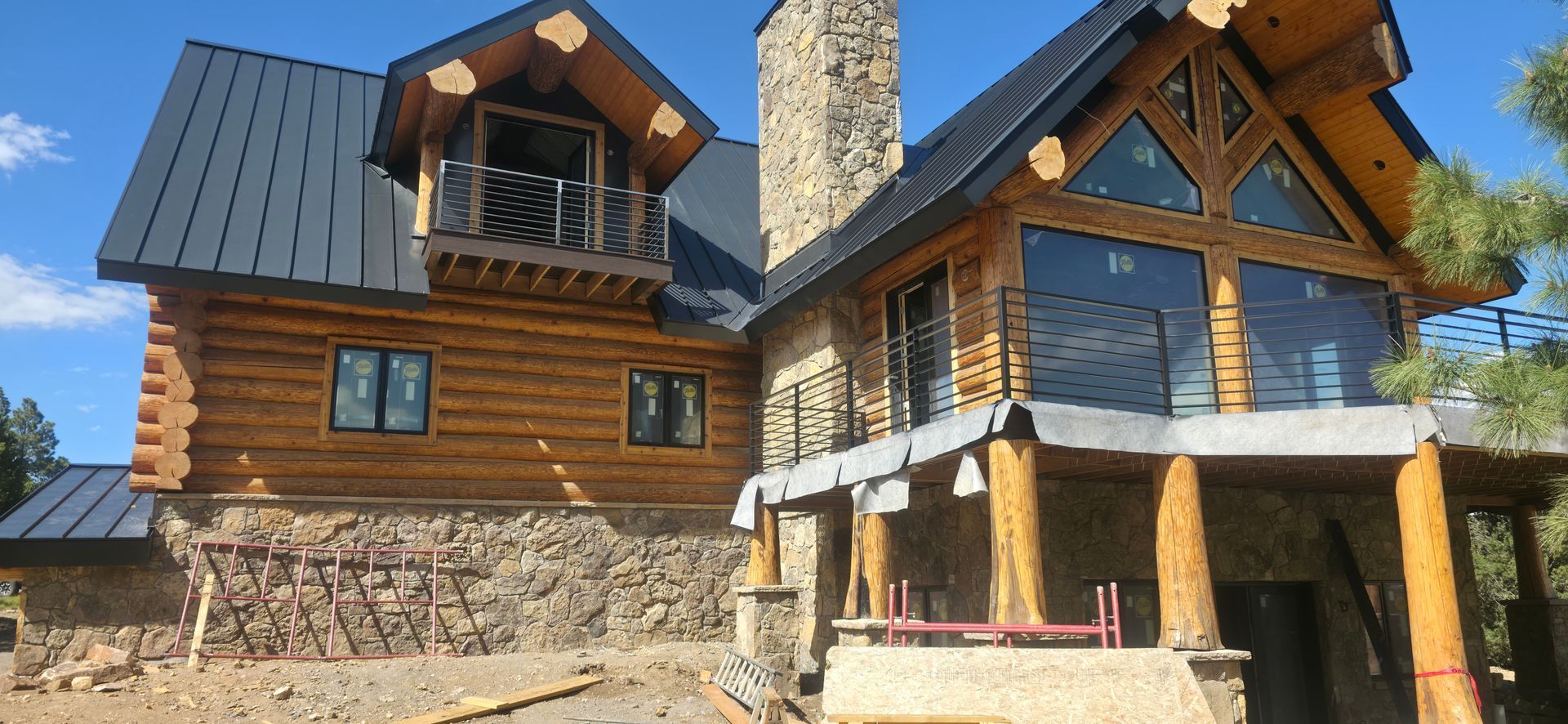 Wooden log house under construction with a stone porch and blue metal roof under a clear sky