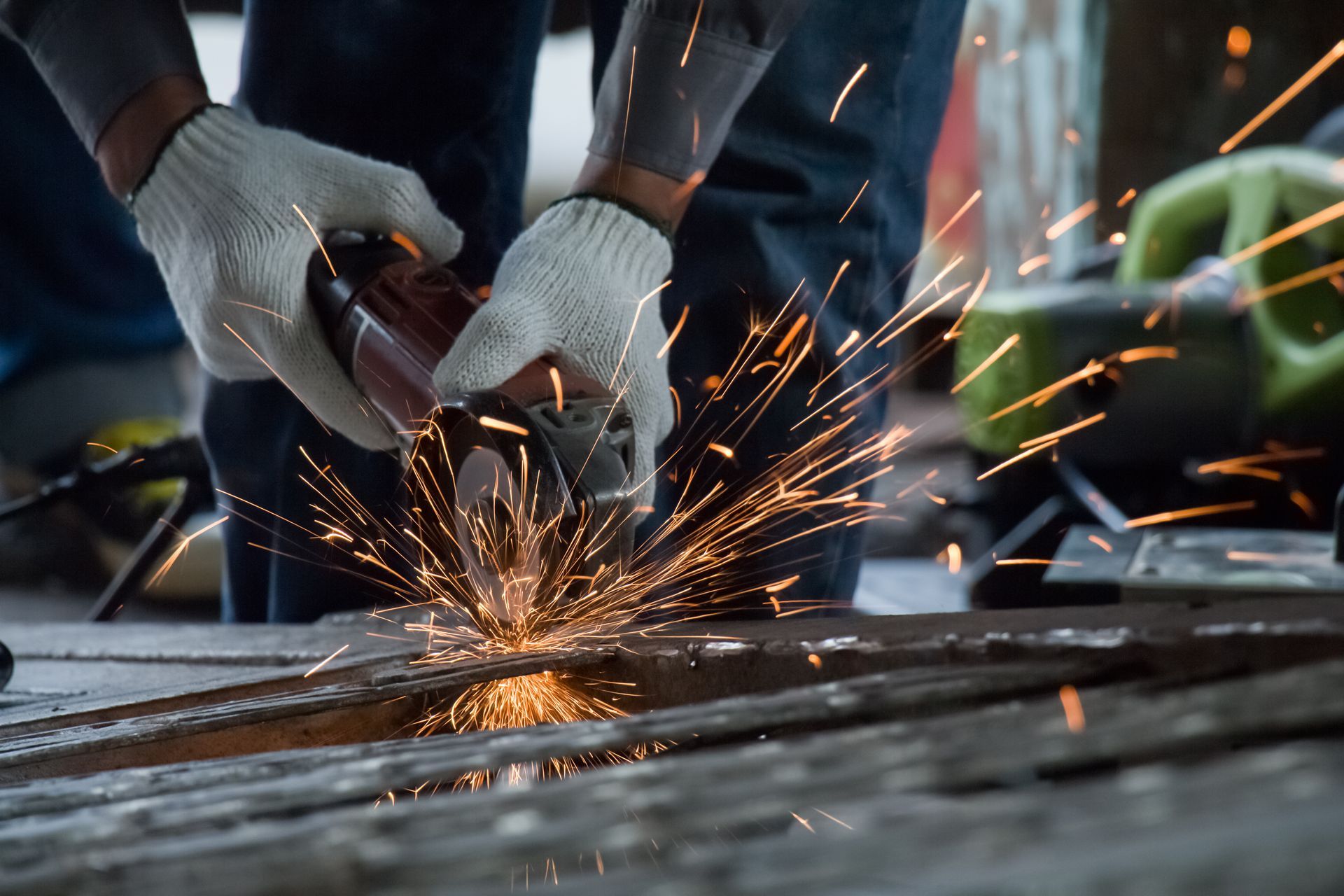 Gloved hands using a grinder on metal, sending bright sparks across a workshop floor.