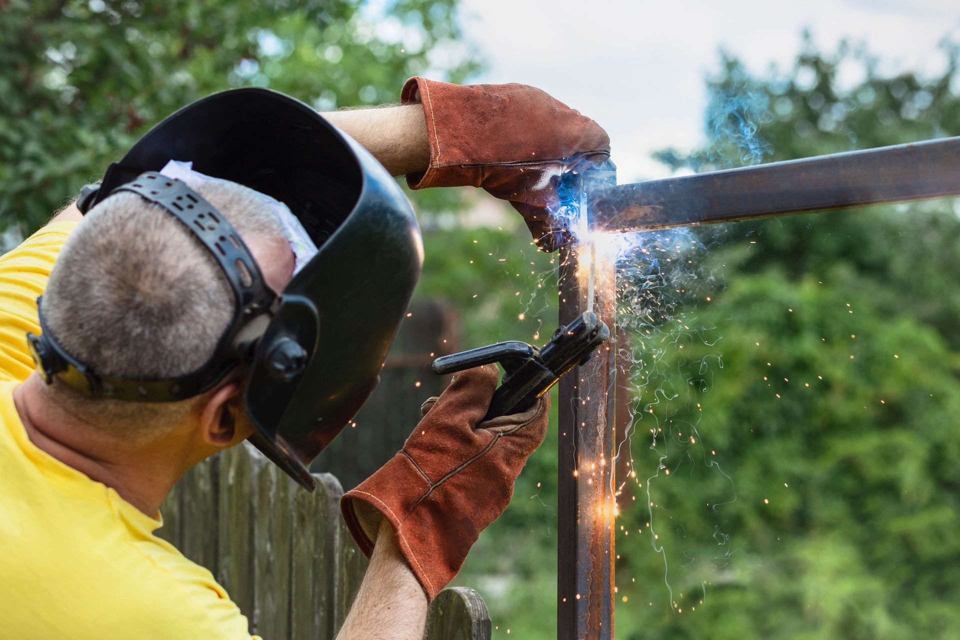 Person welding a metal fence post outdoors, with sparks flying and protective gear on