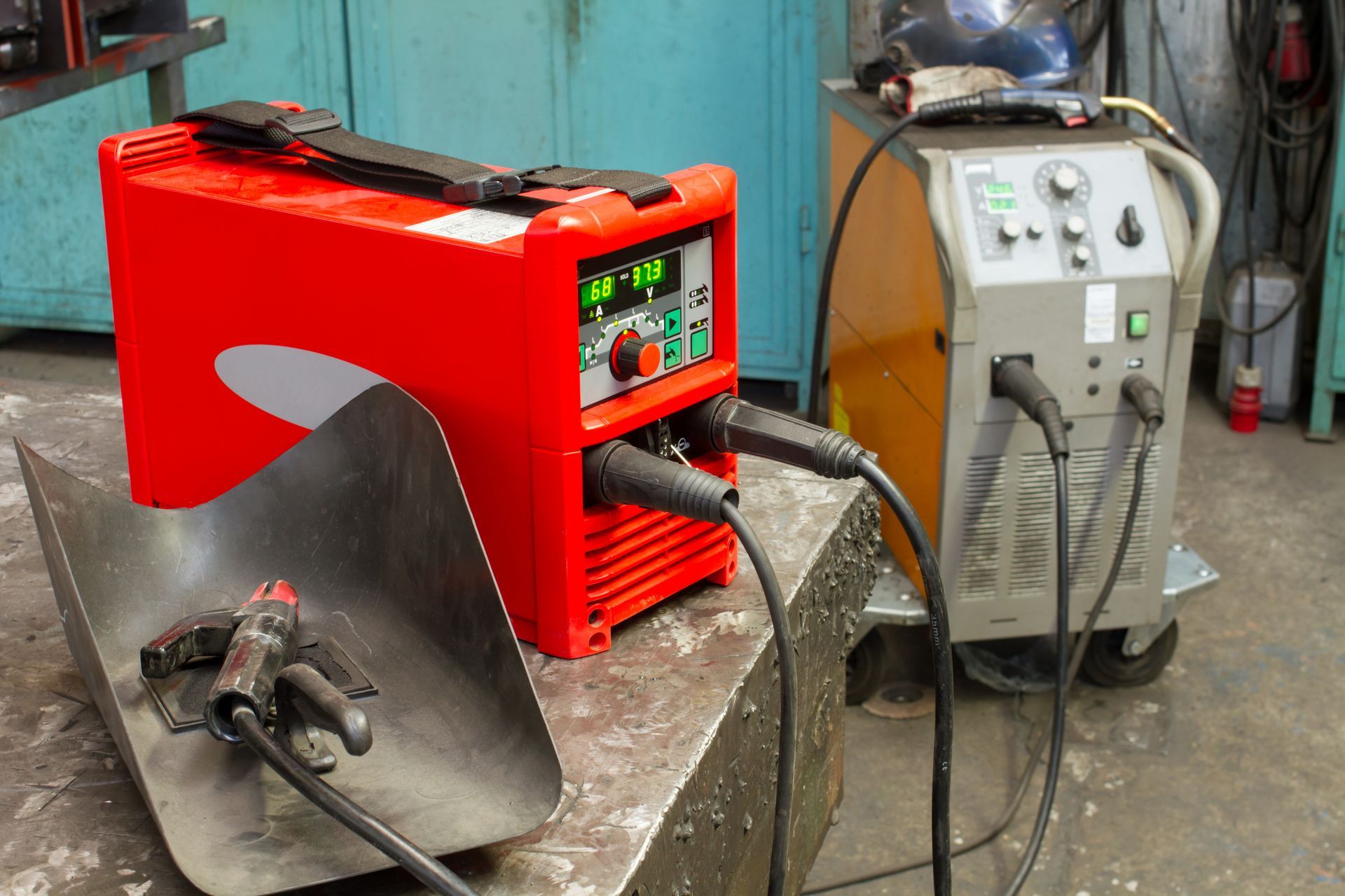 Red welding machines and cables in a workshop, with a metal sheet and torch in the foreground
