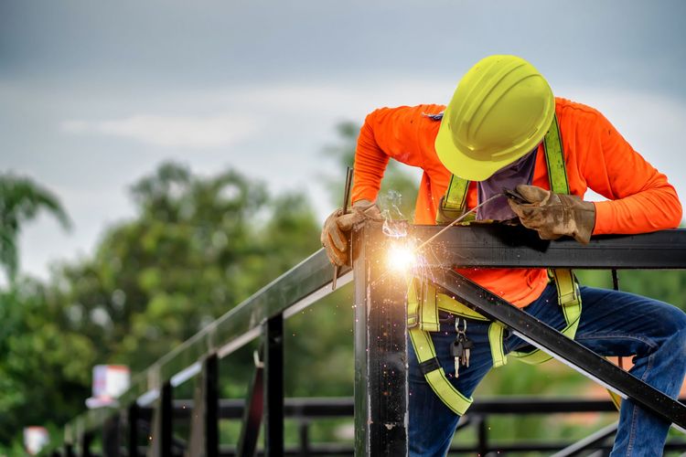 Construction worker in orange safety gear welding a metal railing outdoors with bright sparks