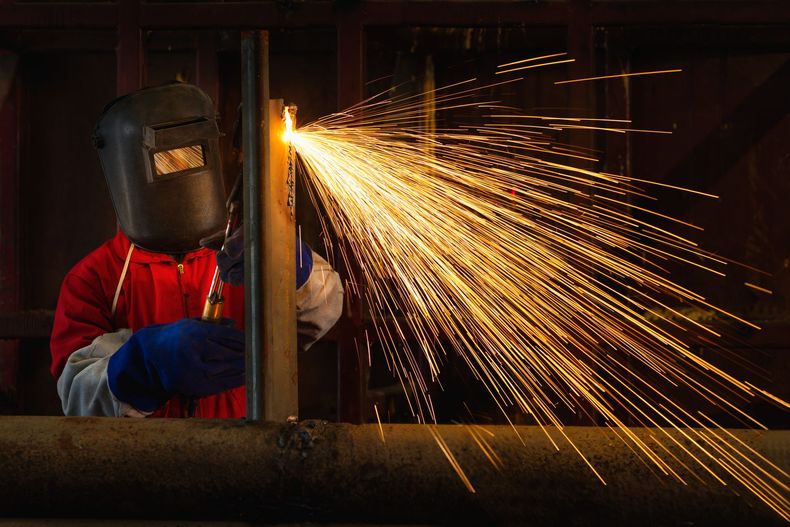 Welder in red shirt and face shield cuts metal, sending bright sparks across a dark workshop