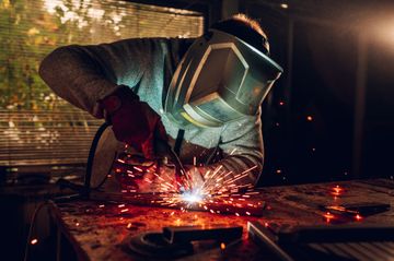 Person welding metal indoors, sparks flying from the workpiece in a dim workshop.