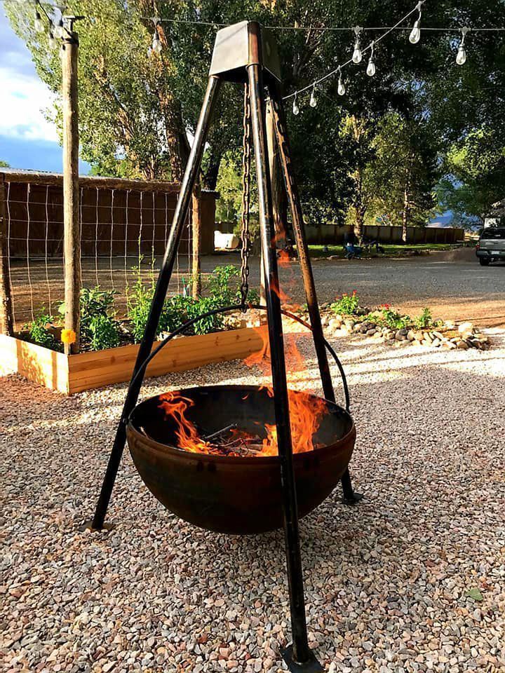 Tripod fire pit with a burning bowl in a gravel courtyard under trees
