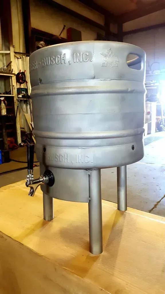Large silver metal tank or vat on four legs in a workshop, with valves and piping attached