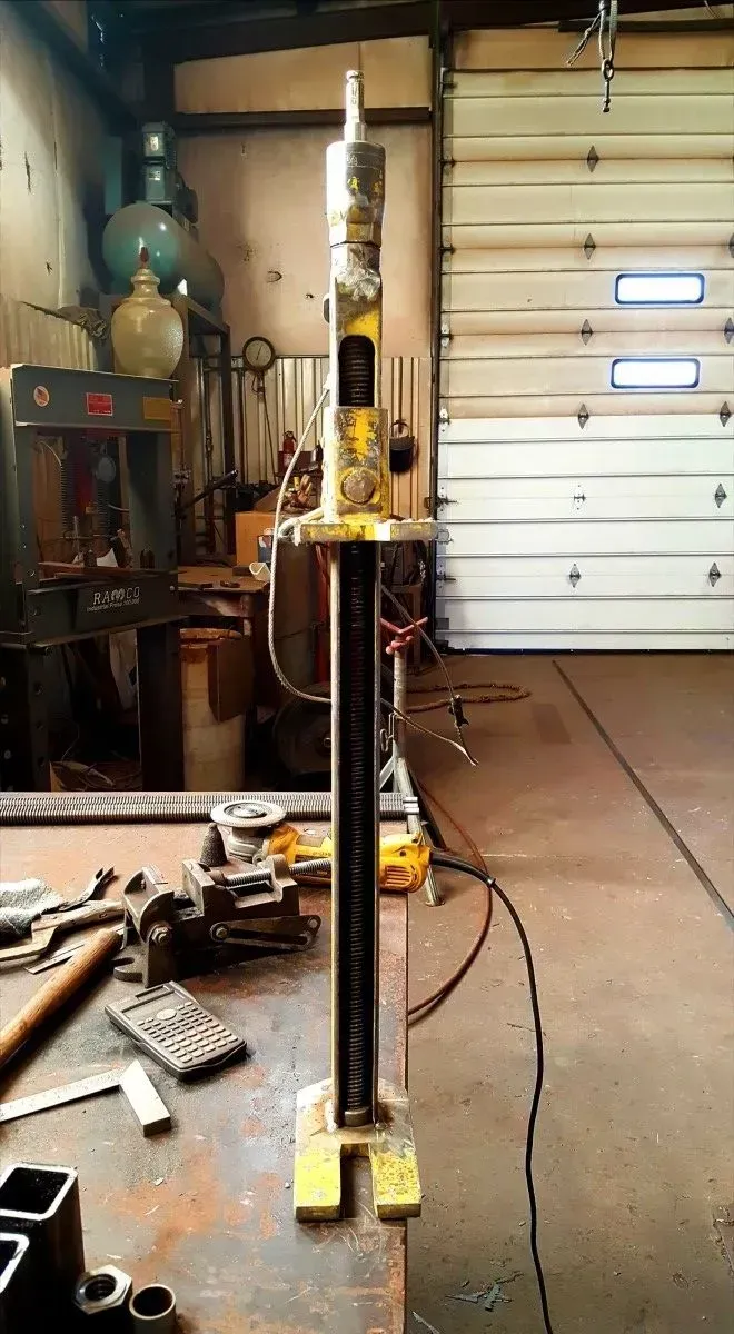 Workshop with a tall black metal post mounted on yellow blocks, tools scattered on a workbench nearby.