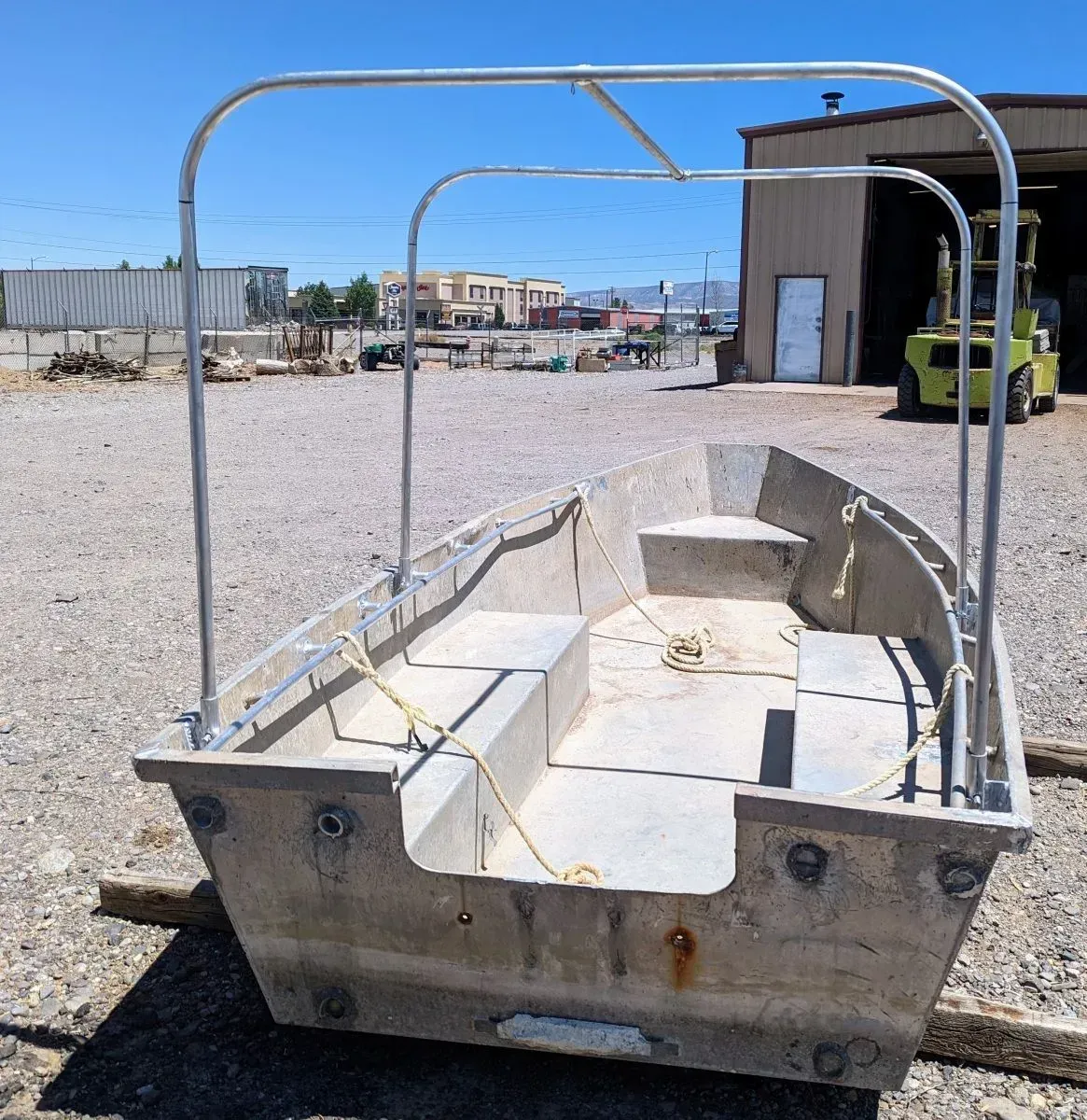 Small white boat on a trailer in a gravel yard near a warehouse, with metal canopy frame overhead.