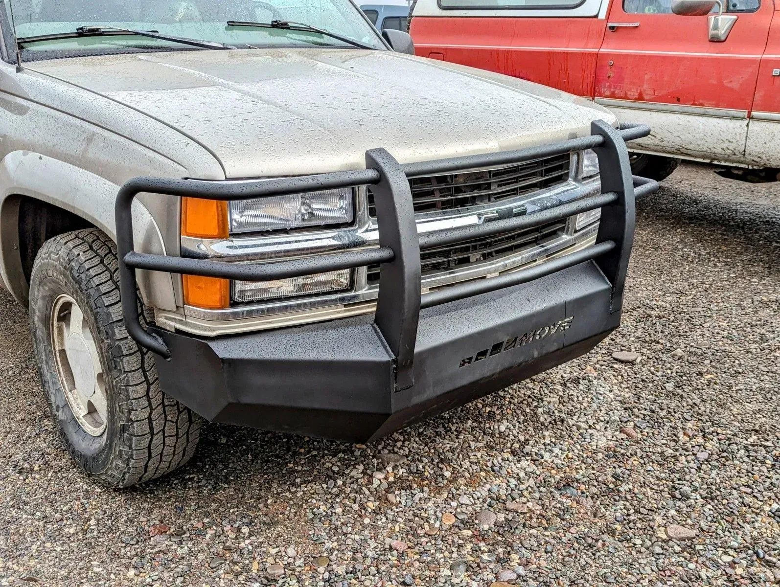 Gray Chevrolet pickup truck with a black grille guard parked on gravel next to a red truck