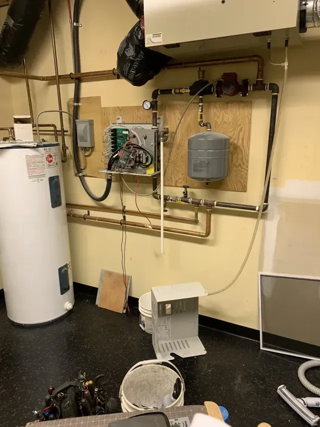 Water heater and heating system components mounted on a yellow wall in a basement setting.