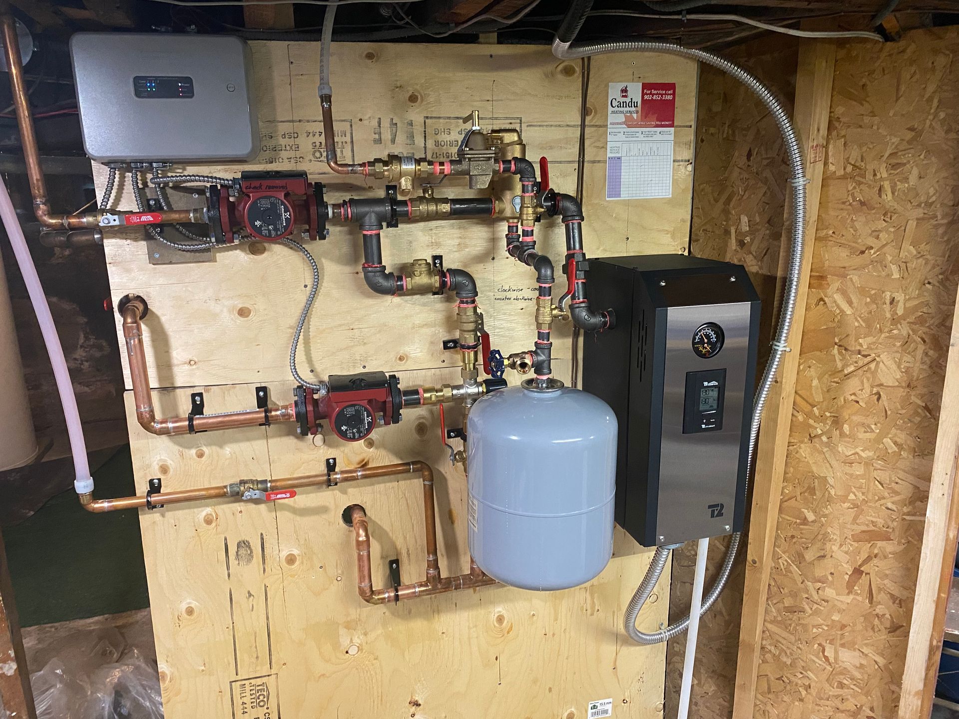 Heating system with copper pipes, pumps, and a water tank mounted on wood paneling.