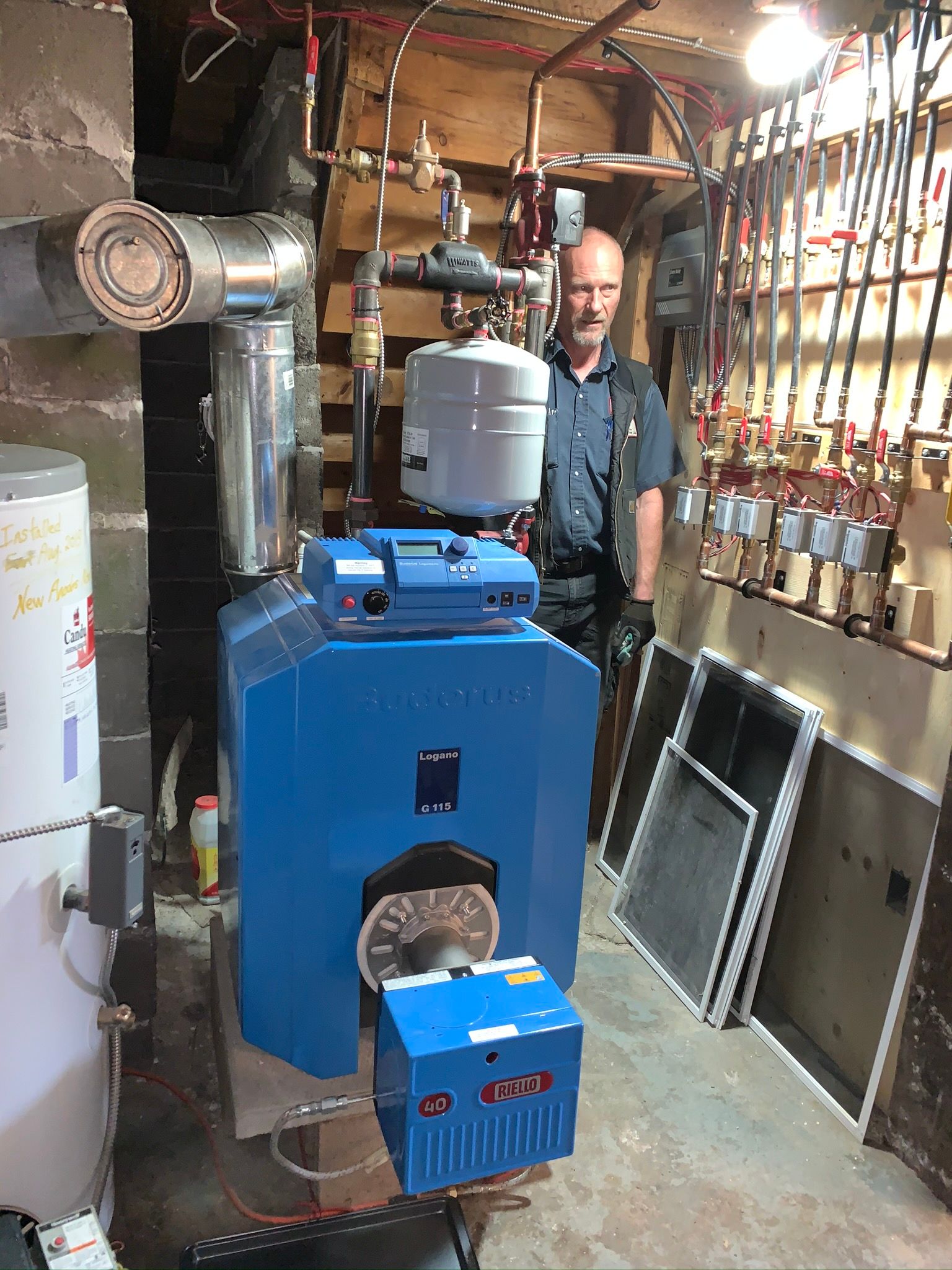 Man standing near a blue boiler in a utility room; copper pipes and tank visible.