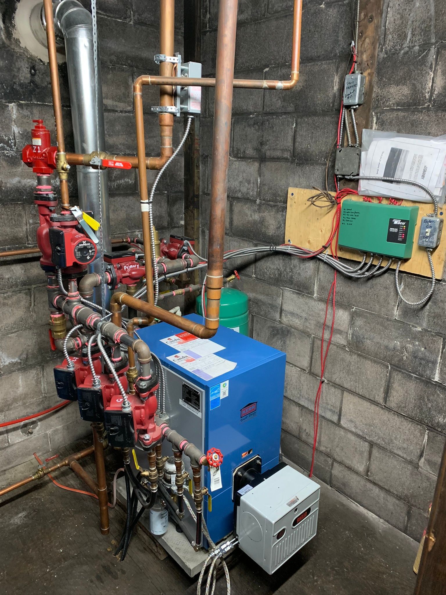 A blue boiler is sitting in a basement next to a brick wall.